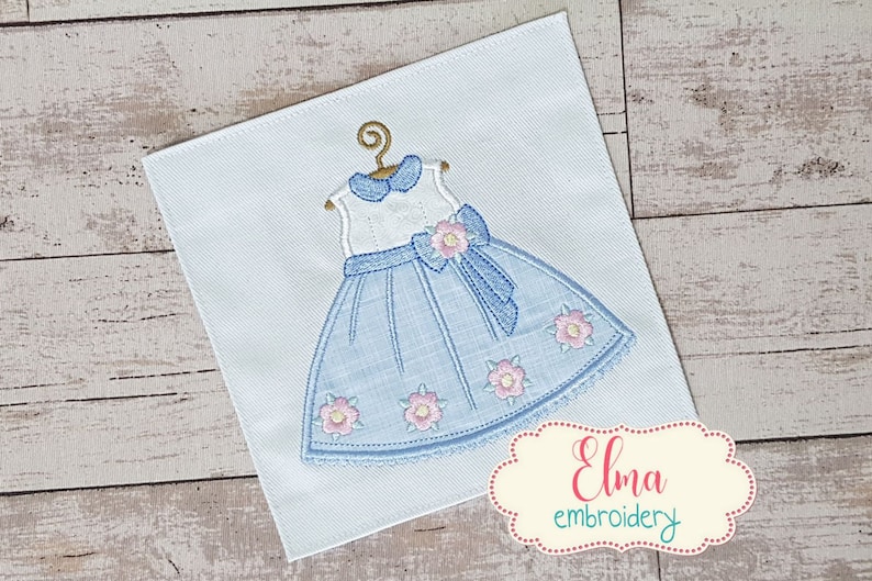 May include: Embroidered design of a blue dress with a white bodice, a pink bow, and pink flowers. The design is on a white fabric background.