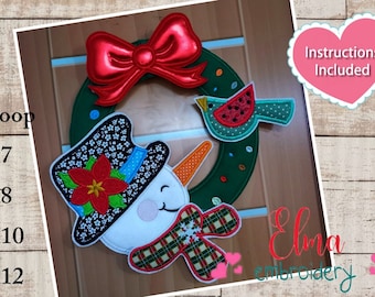 May include: A green wreath with a snowman, a red bow, a poinsettia, and a bird with a red watermelon. The wreath is decorated with red and white plaid fabric, a snowflake, and a blue ribbon. The text "Instructions Included" is in a pink heart shape.