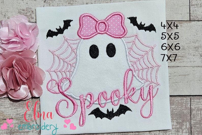 Peut inclure: Tissu blanc brod&eacute; avec un motif de fant&ocirc;me rose. Le fant&ocirc;me a des yeux noirs, un n&oelig;ud rose et est entour&eacute; d'une toile d'araign&eacute;e. Le mot "Spooky" est &eacute;crit en rose en dessous, avec des chauves-souris au-dessus et en dessous. Le texte "4X4, 5X5, 6X6, 7X7" est &agrave; droite.