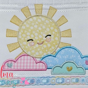 May include: Embroidered design of a smiling yellow sun with a pink and white checkered cloud, a blue cloud with white floral pattern, and a light green cloud with a yellow heart. The design is on a white background with a white lace border.