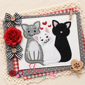 Puede incluir: Una pieza de tela decorativa con tres gatos bordados en gris, blanco y negro. El diseño incluye corazones rojos, lazos, una rosa roja y el texto "Elma emley". La pieza está sobre un fondo a cuadros gris y rojo con ribete de encaje.