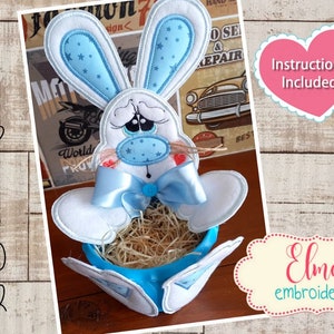 Könnte beinhalten: Ein blauer und weißer Osterhase mit einer blauen Schleife sitzt in einem blauen Osterkorb, der mit Stroh gefüllt ist. Der Hase hält ein kleines blaues Herz. Das Bild enthält den Text "Hoop 5x7 5x8 6x10 7x12" und "Instructions Included".