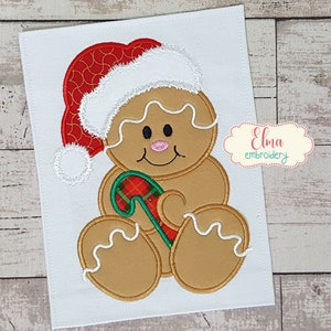 Puede incluir: Una persona de jengibre con un gorro de Papá Noel rojo y blanco y sosteniendo un bastón de caramelo. La persona de jengibre está sentada con las piernas cruzadas. El fondo es una tela blanca.