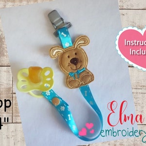 May include: A blue and white ribbon pacifier clip with a brown embroidered dog design. The clip is attached to a pacifier and has a metal clip. The ribbon has a bone and paw print design. The text "Instructions Included" is on a pink heart. The text "Hoop 4x4" is on a wooden surface. The text "Elma embroidery" is on a white surface.