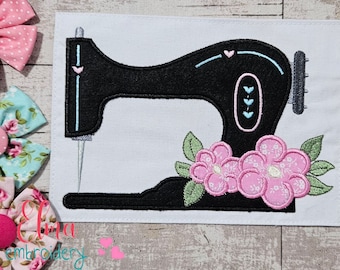 May include: Black sewing machine applique with pink and green floral accents on a white background. The sewing machine has light blue and pink details. The image is on a white background with a wooden surface.