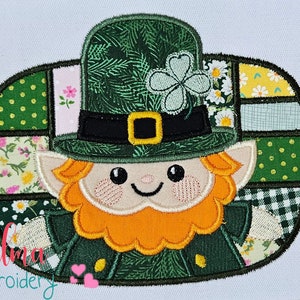 May include: A green and orange patchwork appliqué of a leprechaun wearing a green hat with a shamrock on top. The leprechaun has a red beard and a happy face. The design is available in sizes 4x4, 5x4, 5x7, 5x8, 6x10, and 7x12 inches.