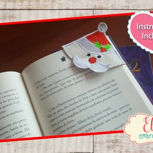 K&ouml;nnte beinhalten: Ein wei&szlig;-roter Weihnachtsmann-Lesezeichen mit einem gr&uuml;nen Stechpalmenzweig. Das Lesezeichen liegt auf einem Buch mit dem Text "Instructions Included" in einem rosa Herz. Das Lesezeichen ist f&uuml;r einen 5x7-Hoop.