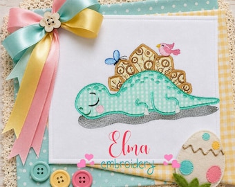 Könnte beinhalten: Ein weißes Stoffquadrat mit einem gestickten mintgrünen Dinosaurier mit gemustertem Rücken, einem blauen Schmetterling und einem rosa Vogel. Der Text Elma embroidery ist darunter gestickt. Eine bunte Satinschleife und ein Filzei sind befestigt.