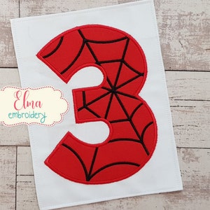 May include: A red number 3 applique with a black web design, embroidered on a white fabric. The number is a bold, eye-catching design. The image also includes the text "Elma embroidery".