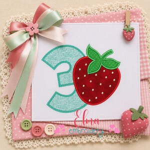 May include: A white fabric square featuring an embroidered teal number three and a red strawberry with green leaves. A large pink and green ribbon bow is attached to the top left. A small strawberry clip and three buttons are included as accents.