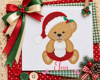 May include: A white fabric square featuring an embroidered tan teddy bear wearing a red polka-dot Santa hat and a white apron. The bear is accented with holly leaves and red berries. The name Elma is embroidered in red script below the bear.