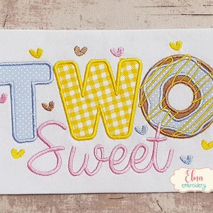 Könnte beinhalten: Stickerei mit dem Wort "ZWEI" in Gelb und Blau mit einem Gingham-Muster auf dem "W". Das Wort "Sweet" ist darunter in rosa Kursivschrift gestickt. Ein Donut mit gelber Glasur und Streuseln befindet sich rechts neben dem Wort "ZWEI".