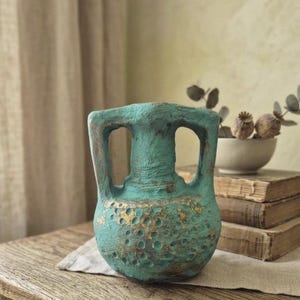 Op de afbeelding: Een turquoise vaas in amfoorstijl met een getextureerd, verweerd oppervlak met goudkleurige accenten en inkepingen. De vaas heeft twee opvallende handvatten aan de zijkant, een brede buik en een smalle hals met een rustieke uitstraling.