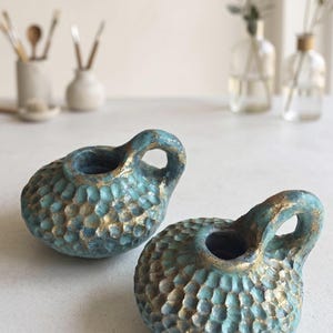 Pode incluir: Dois pequenos vasos de cerâmica turquesa com superfície texturizada e detalhes dourados. Cada vaso tem uma alça em forma de laço. Os vasos estão sobre uma superfície branca, com outros objetos decorativos ao fundo.