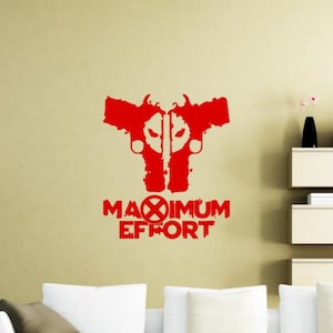Peut inclure: Un autocollant mural rouge représentant un graphique symétrique de deux pistolets orientés vers l'extérieur avec une forme de masque stylisée au centre. Sous le graphique, le texte indique MAXIMUM EFFORT, avec le X remplacé par un réticule.