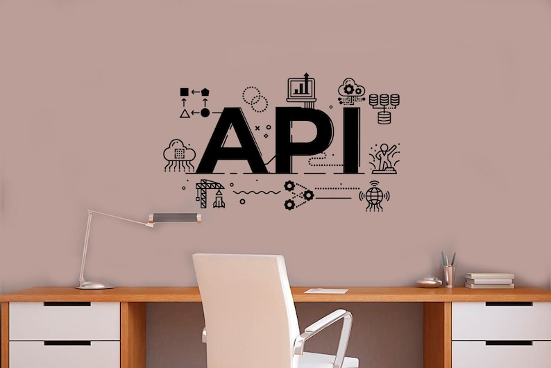 Op de afbeelding: Een zwart-witte illustratie van het woord "API" met verschillende pictogrammen en symbolen eromheen, waaronder een wolk, een computer, tandwielen en een wereldbol. De illustratie bevindt zich op een lichtroze muur achter een wit bureau met een witte stoel.