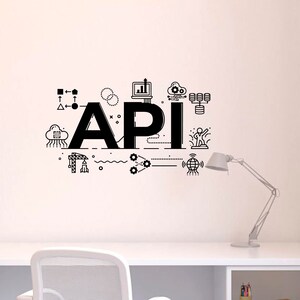 Op de afbeelding: Een zwart-witte illustratie van het woord "API" met verschillende pictogrammen en symbolen eromheen, waaronder een wolk, een computer, een wereldbol en een raket. De illustratie bevindt zich op een witte muur boven een wit bureau met een witte stoel en een lamp.