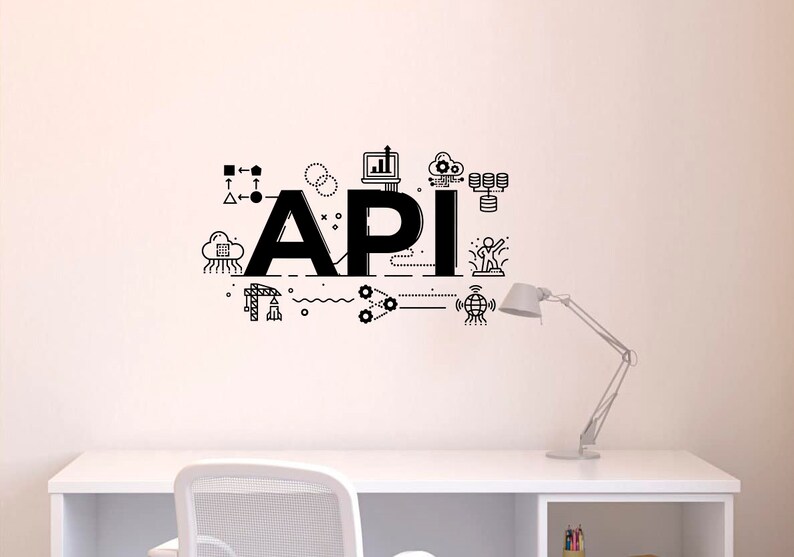 Op de afbeelding: Een zwart-witte illustratie van het woord "API" met verschillende pictogrammen en symbolen eromheen, waaronder een wolk, een computer, een wereldbol en een raket. De illustratie bevindt zich op een witte muur boven een wit bureau met een witte stoel en een lamp.