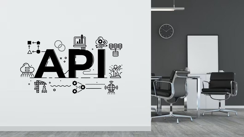 Op de afbeelding: Een witte muur met een zwart-witte illustratie van het woord "API" omringd door verschillende pictogrammen en symbolen. De illustratie is in een moderne, minimalistische stijl. De muur bevindt zich in een kantooromgeving met twee zwarte bureaustoelen en een klok aan de muur.