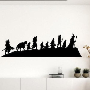Op de afbeelding: Zwarte silhouet muursticker met een groep figuren, waaronder een tovenaar, boogschutters en ruiters te paard, die door een landschap lopen. De figuren staan tegen een witte muur, wat een opvallend contrast cre&euml;ert.