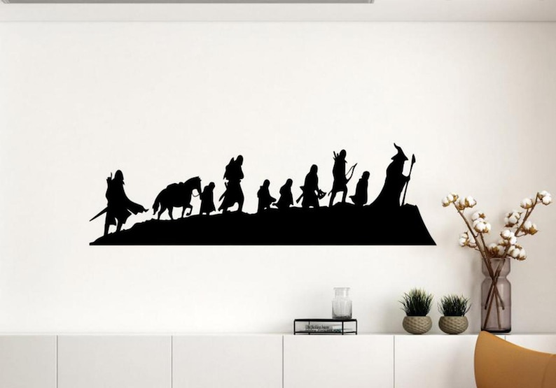 Op de afbeelding: Zwarte silhouet muursticker met een groep figuren, waaronder een tovenaar, boogschutters en ruiters te paard, die door een landschap lopen. De figuren staan tegen een witte muur, wat een opvallend contrast cre&euml;ert.
