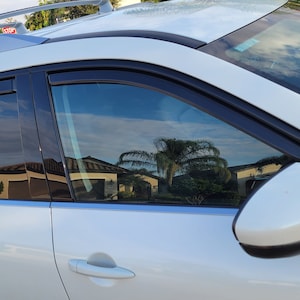 May include: A white SUV with black window visors and a roof rack. The windows reflect a cloudy sky and houses. The side mirror is white and black. The door handle is visible.