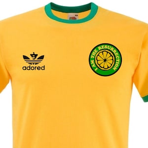 May include: A yellow t-shirt with a green ringer. The shirt features a black Adidas logo with the word "adored" underneath. The shirt also has a green circle with a lemon inside and the text "I am the resurrection" around it.