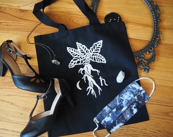 May include: Black tote bag with a white graphic of a stylized tree with a human-like figure and the text "C/th". The bag is on a wooden surface with a pair of black shoes, a necklace, a small white stone, and a blue and white patterned face mask.