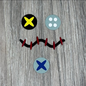 May include: A wooden surface with a stitched mouth, two black buttons with yellow and blue crosses, and a gray button with three white dots.