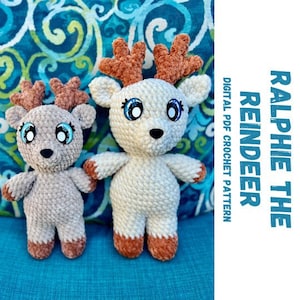 May include: Two crocheted reindeer toys, one brown and white and one beige. The brown and white reindeer has blue eyes and a brown nose. The beige reindeer has blue eyes and a brown nose. The toys are sitting on a blue and green patterned surface. The text "Ralphie the Reindeer" is in the background.