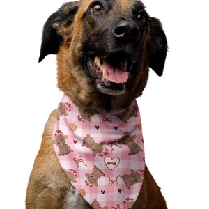 May include: A happy dog wearing a pink and white checkered bandana. The bandana features a repeating pattern of brown teddy bears, red hearts, and bows. The dog has brown and black fur and is smiling with its tongue out.