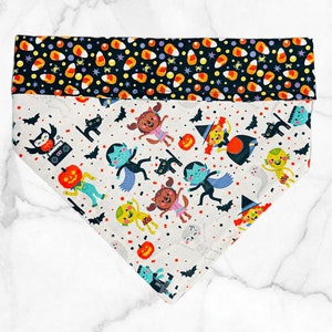 May include: A triangular pet bandana featuring a top border of candy corn and stars on a black background. The main section is off-white with a repeating pattern of cartoon monsters, pumpkins, bats, and black cats in vibrant colors.