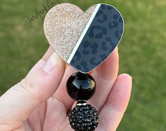 May include: A black and gold glitter heart-shaped badge holder with a leopard print design. The holder is attached to a black beaded retractable badge reel.