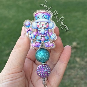 May include: A teal and purple glitter badge reel with a nutcracker design. The nutcracker is wearing a pink and blue uniform with a crown and holding a candy cane.