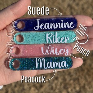 Puede incluir: Cinco llaveros de acr&iacute;lico transparente con purpurina de diferentes colores. Los llaveros est&aacute;n etiquetados con los nombres "Jeannine", "Riker", "Wifey", "Mama" y los colores "Suede", "Thin Mint", "Peach" y "Peacock".