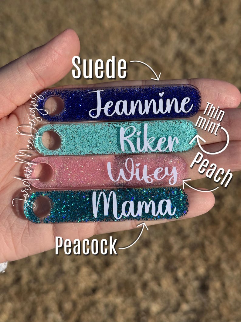Puede incluir: Cinco llaveros de acr&iacute;lico transparente con purpurina de diferentes colores. Los llaveros est&aacute;n etiquetados con los nombres "Jeannine", "Riker", "Wifey", "Mama" y los colores "Suede", "Thin Mint", "Peach" y "Peacock".
