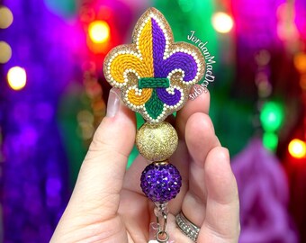 May include: A Mardi Gras-themed badge reel featuring a fleur-de-lis design in gold, yellow, purple, and green glitter. The badge reel has a gold glitter bead and a purple rhinestone bead. The text "JordanMacDesigns" is visible.