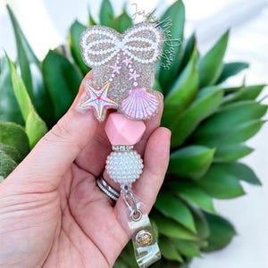 May include: A badge reel with a glittery bow, starfish, and seashell design. The badge reel has a pink heart and pearl bead accents. The reel is attached to a clear plastic clip.