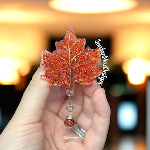 May include: A maple leaf badge reel featuring a vibrant orange and red glitter finish with a gold-toned vein detail. The badge reel includes a retractable cord mechanism and a small circular accent piece with matching orange glitter at the base.