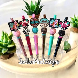 Puede incluir: Una colección de seis bolígrafos decorativos con varios diseños. Cada bolígrafo presenta un barril de diferente color, una sección de cuentas y un dije con la palabra "Mama" y detalles florales. Los bolígrafos se exhiben en un soporte de plástico transparente.