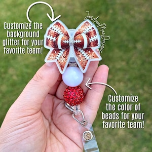Puede incluir: Un portatarjetas con un adorno en forma de lazo con patrón de fútbol americano y purpurina cobriza, acentuado por una cuenta blanca y una cuenta roja de pedrería. El texto dice: Customize the background glitter for your favorite team! Customize the color of beads for your favorite team!