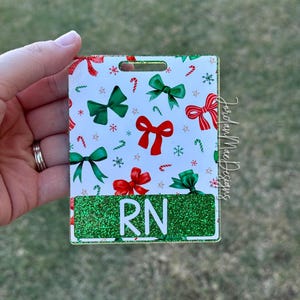 Puede incluir: Insignia de brillo verde con fondo blanco y un patrón de lazo navideño. La insignia tiene una ventana de plástico transparente con las letras "RN" en blanco.