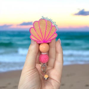 May include: A badge reel featuring a glittery gold seashell topper with a bright pink outline and the text JordanMacDesigns. The topper is attached to a vertical stack of beads including a smooth peach bead, a small white bead, and a textured pink bead.