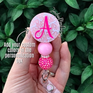 May include: A pink and white retractable badge reel with a large pink letter "A" on the top. The badge reel has a pink bead and a sparkly pink bead. The text "Add your custom colors in the personalization box" is in the image.