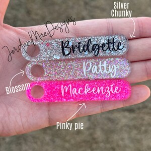 Puede incluir: Tres llaveros personalizados con purpurina incrustada en el pl&aacute;stico transparente. El llavero superior es de purpurina plateada con el nombre "Bridgette" escrito en negro. El llavero del medio es de purpurina rosa con el nombre "Patty" escrito en negro. El llavero inferior es de purpurina rosa intenso con el nombre "Mackenzie" escrito en negro.