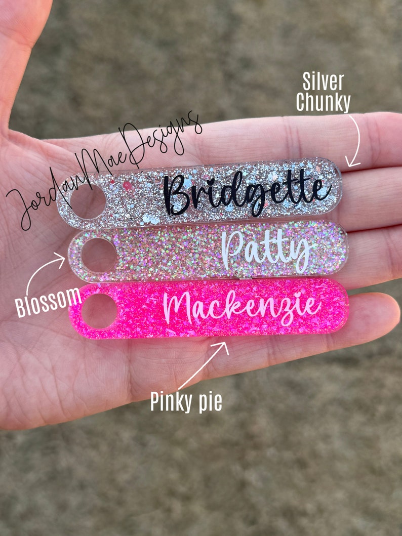 Puede incluir: Tres llaveros personalizados con purpurina incrustada en el pl&aacute;stico transparente. El llavero superior es de purpurina plateada con el nombre "Bridgette" escrito en negro. El llavero del medio es de purpurina rosa con el nombre "Patty" escrito en negro. El llavero inferior es de purpurina rosa intenso con el nombre "Mackenzie" escrito en negro.