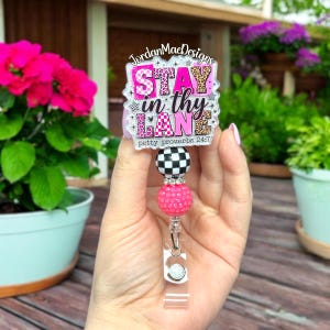 May include: A badge reel featuring a rectangular acrylic topper with the text JordanMaeDesigns and Stay in thy LANE petty proverbs 24:7 in pink and leopard print. The reel includes a black and white checkered bead and a textured pink bead.