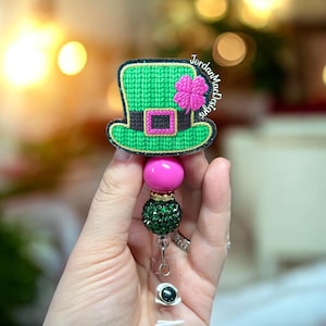 Puede incluir: Un portatarjetas con temática del Día de San Patricio, verde y negro, con un diseño de sombrero de duende y un trébol rosa. El portatarjetas tiene una cuenta rosa, una cuenta de purpurina verde y un cordón retráctil transparente. El texto "JordanMacDesigns" es visible.