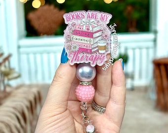 Può includere: Un porta badge rosa e bianco con la scritta "Books Are My Therapy". Il porta badge presenta una pila di libri, fiori e una tazza di caffè. Il porta badge ha una perla argentata, una perla rosa strutturata e una clip di plastica trasparente.