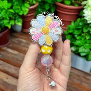 May include: A daisy-shaped badge reel with a glittery silver border. The flower has blue gingham, pink plaid, and yellow polka dot petals. Below the flower are yellow and iridescent pink beads. The badge reel has a clear plastic clip and a silver button.